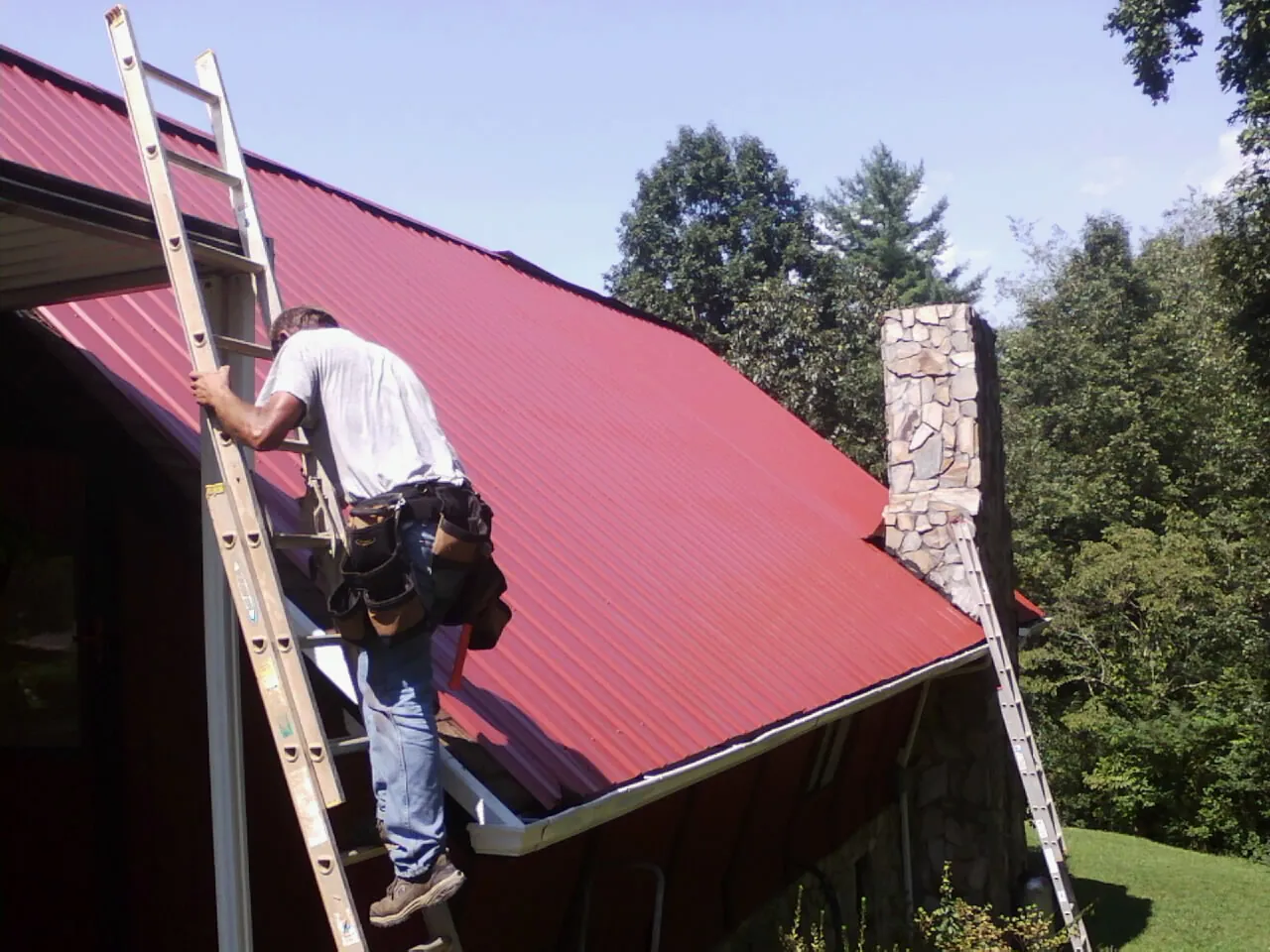 Roofer on a ladder beside a red metal roof and stone chimney — Soffit & Fascia Repair by Roofing Brothers in Rush City